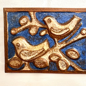 May include: A ceramic wall tile with a blue background and two white birds perched on a brown branch. The birds are stylized and have a simple design. The tile is surrounded by a brown frame.