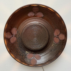 May include: A large, brown ceramic plate with a raised, concentric circle design in the centre. The plate is decorated with four stylised leaf designs in shades of brown and pink.