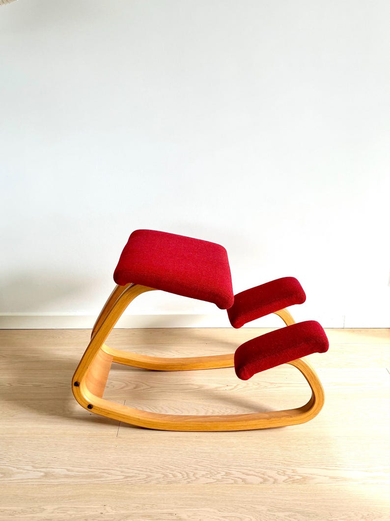 May include: A kneeling chair with a curved, light-coloured wooden frame and red fabric cushions. The chair has a seat and two knee rests, designed for ergonomic support. The chair is on a light wood floor.
