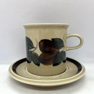 Peut inclure: Une tasse à café et une soucoupe en céramique beige avec un motif floral brun et vert. La tasse a une poignée arrondie et la soucoupe a une bande brune autour du bord.