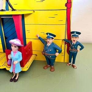 Op de afbeelding: Een kleurrijk speelgoed treinstation in geel met blauwe en rode details, met drie kleine beeldjes. De beeldjes dragen blauwe uniformen en een lichtblauwe jas. De sc&egrave;ne speelt zich af op een groen oppervlak.