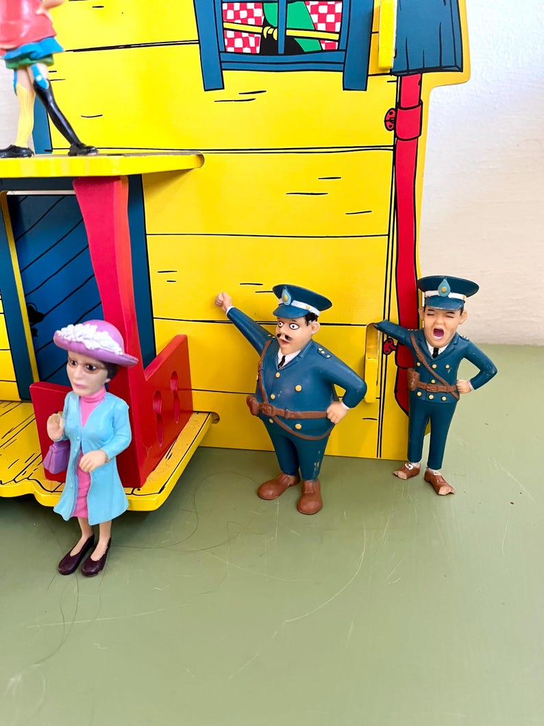 Op de afbeelding: Een kleurrijk speelgoed treinstation in geel met blauwe en rode details, met drie kleine beeldjes. De beeldjes dragen blauwe uniformen en een lichtblauwe jas. De sc&egrave;ne speelt zich af op een groen oppervlak.