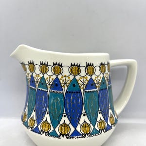 Può includere: Un brocca per la panna in ceramica bianca con un motivo a pesci blu, verdi e dorati. La brocca ha un manico e un corpo arrotondato.