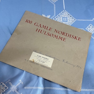 Op de afbeelding: Een bruin papieren boekje met de titel "100 GAMLE NORDISKE HULSØMME" in rode letters. Het boekje is gelabeld "Kristine Frederiksen. Ferlumrej 27. a" in handschrift. Een wit label met de tekst "CARL PERMIN" en "C.B. GARN" is aan het boekje bevestigd.