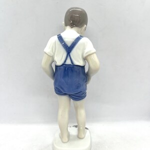 May include: A porcelain figurine of a child standing and facing away from the viewer. The figure wears a white short-sleeved shirt and blue shorts with suspenders that cross at the back. The child has brown hair and is barefoot on a white base.