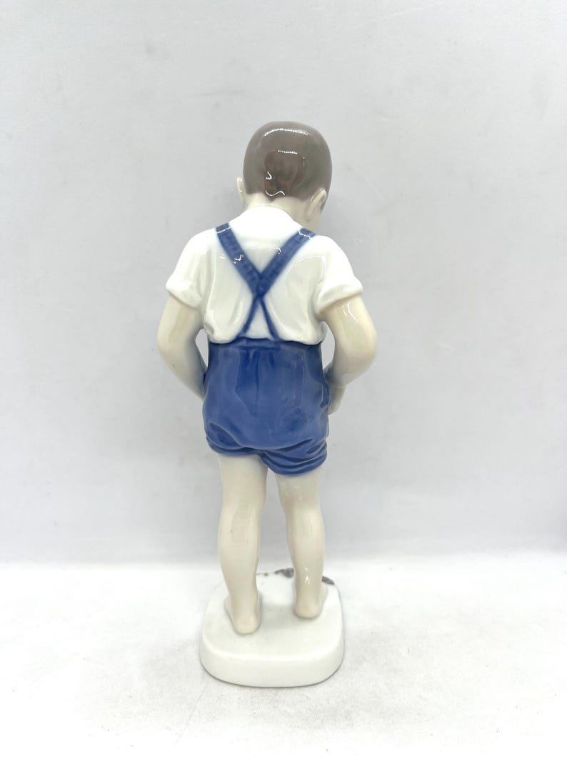 May include: A porcelain figurine of a child standing and facing away from the viewer. The figure wears a white short-sleeved shirt and blue shorts with suspenders that cross at the back. The child has brown hair and is barefoot on a white base.