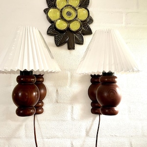 Può includere: Una coppia di lampade da parete con basi in legno tornito marrone scuro e paralumi conici plissettati bianchi, posizionate sotto una placca decorativa in ceramica a forma di fiore giallo con foglie marrone scuro e centro testurizzato.