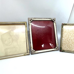 Può includere: Tre cornici rettangolari per foto. La cornice a sinistra è dorata con angoli decorati. La cornice centrale è argentata con un bordo intrecciato e angoli decorativi, contenente un inserto rosso. La cornice a destra è argentata con agrifoglio.