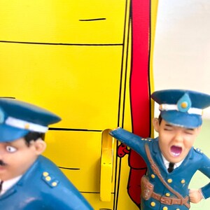 Op de afbeelding: Twee speelgoedagenten in blauwe uniformen. E&eacute;n schreeuwt met open mond. De achtergrond is geel met zwarte lijnen en een rode streep. De figuren zijn in cartoonstijl ontworpen.