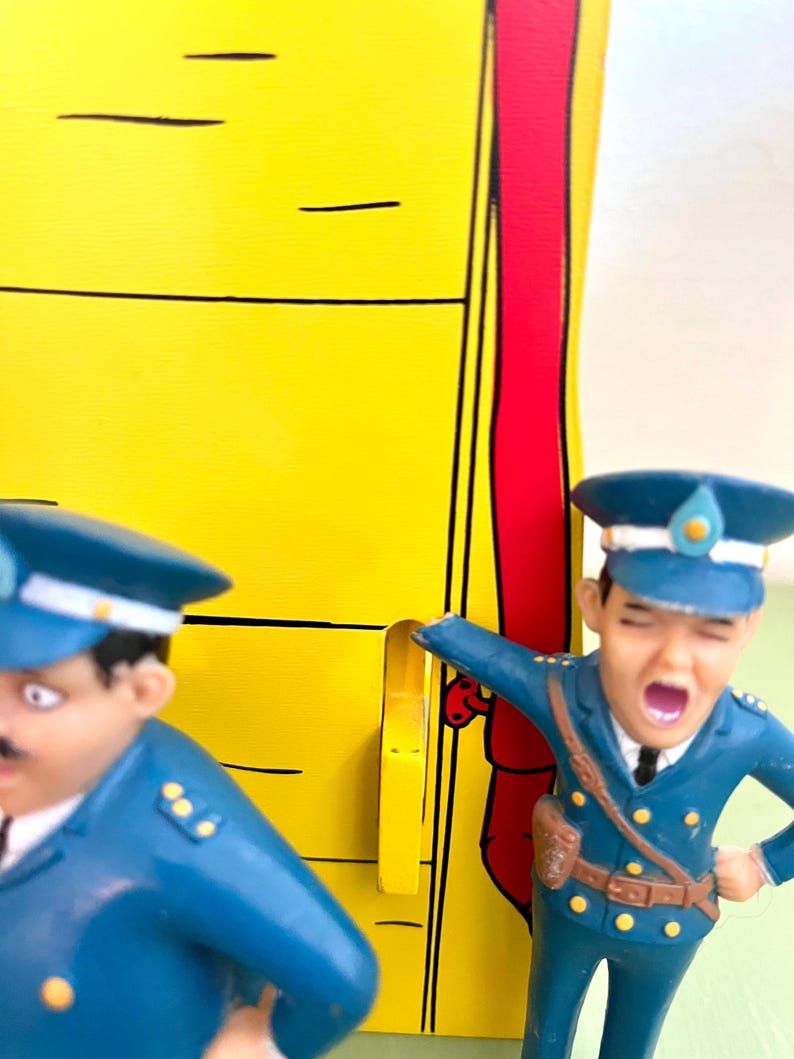 Op de afbeelding: Twee speelgoedagenten in blauwe uniformen. E&eacute;n schreeuwt met open mond. De achtergrond is geel met zwarte lijnen en een rode streep. De figuren zijn in cartoonstijl ontworpen.