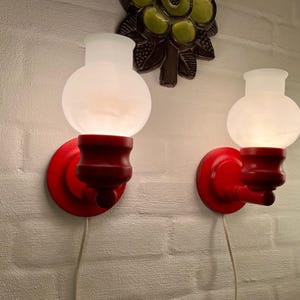 Może przedstawiać: Dwa czerwone kinkiety ścienne z kloszami w kształcie kul z matowego szkła. Kinkiety mają klasyczny design z okrągłą podstawą i ozdobnym trzonem. Są zamontowane na białej ceglanej ścianie. Nad kinkietami znajduje się dekoracja ścienna.