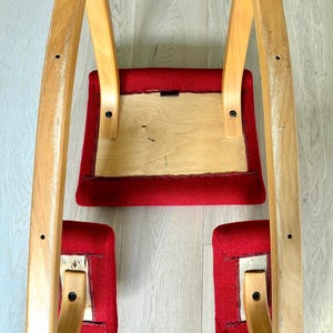 May include: A vintage rocking chair featuring a light wood frame and red fabric seat and footrests. The chair's curved design is visible from an upward angle, showcasing its construction and materials.