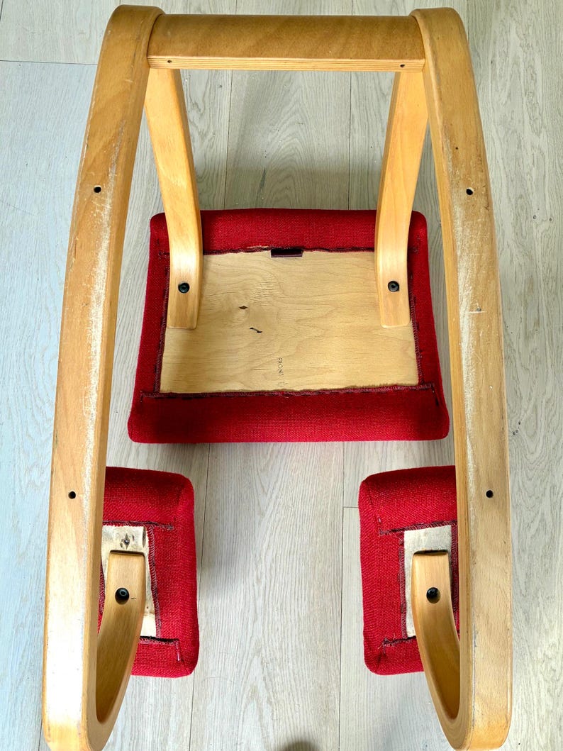 May include: A vintage rocking chair featuring a light wood frame and red fabric seat and footrests. The chair's curved design is visible from an upward angle, showcasing its construction and materials.