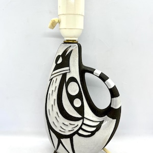 Può includere: Una lampada da tavolo in ceramica bianca con un design di uccello stilizzato in bianco e nero. La lampada ha un cavo bianco e un paralume bianco.