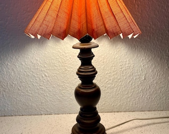 May include: A vintage table lamp with a dark brown, turned wood base and a pleated orange lampshade. The shade is cone-shaped and emits a warm glow. The lamp is on a white surface.