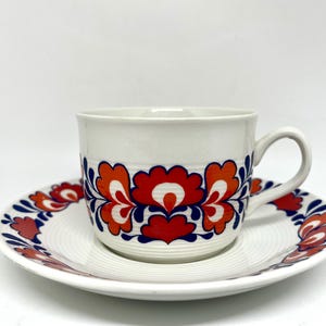 Puede incluir: Juego de taza y platillo de té de cerámica blanca. La taza y el platillo están decorados con un patrón floral repetido en rojo, naranja y azul. El platillo tiene una textura acanalada. Un diseño clásico para servir té o café.