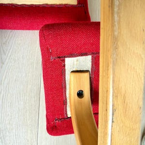 May include: Close-up of a red upholstered chair seat with a wooden frame. The seat is a vibrant red, and the wooden frame is a light, natural colour. The chair appears to be well-constructed and sturdy.