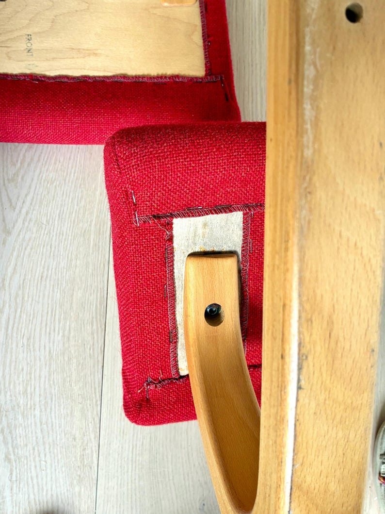 May include: Close-up of a red upholstered chair seat with a wooden frame. The seat is a vibrant red, and the wooden frame is a light, natural colour. The chair appears to be well-constructed and sturdy.