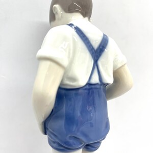 May include: A porcelain figurine of a child viewed from behind, wearing a white short-sleeved shirt and blue overalls with crossed straps. The figure has smooth, glossy skin tones and brown hair, captured in a standing position against a plain background.