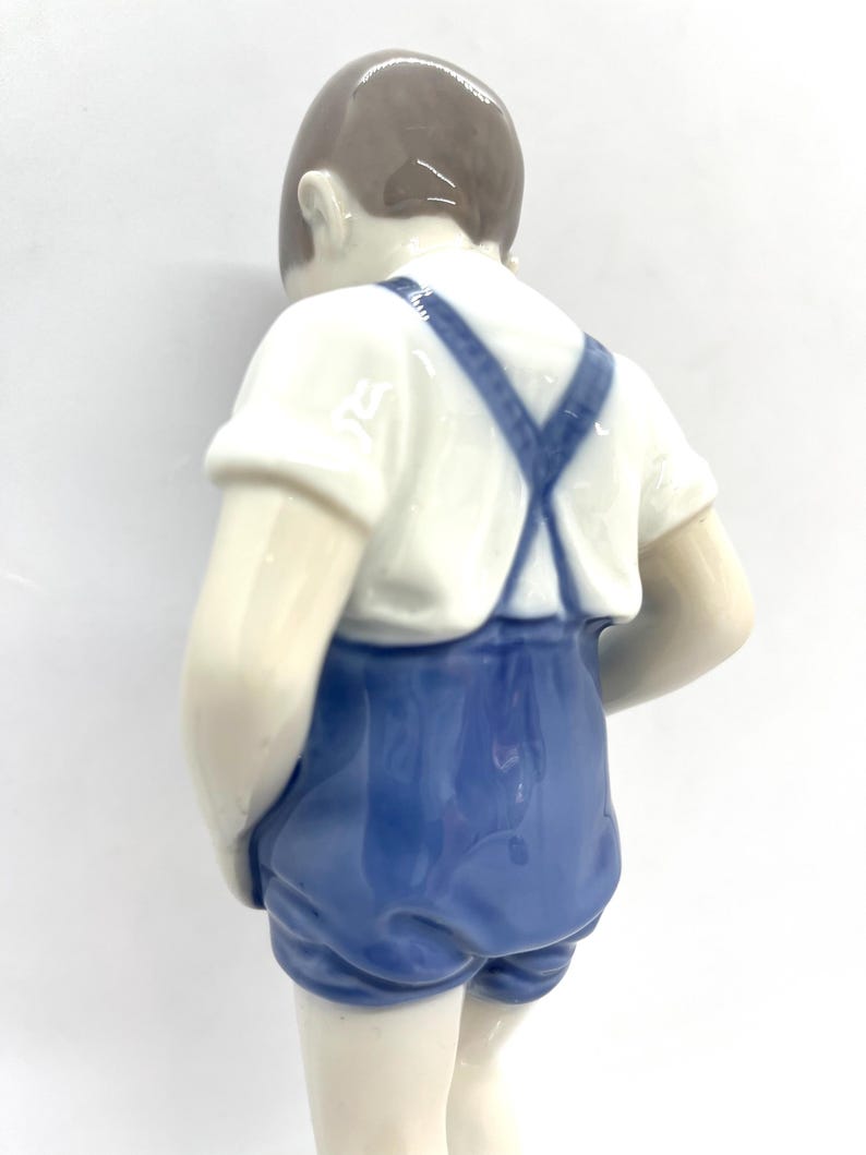 May include: A porcelain figurine of a child viewed from behind, wearing a white short-sleeved shirt and blue overalls with crossed straps. The figure has smooth, glossy skin tones and brown hair, captured in a standing position against a plain background.