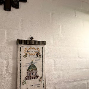 May include: A white embroidered wall hanging with a gold frame. The embroidery depicts a church building with the text "Tårne så tinder" and "Vor Frelsers Kirke" in Danish.
