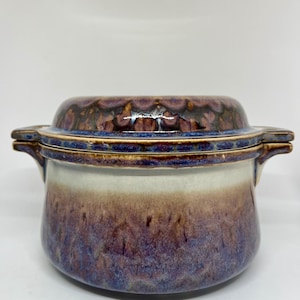 Könnte beinhalten: Eine Keramik-Auflaufform mit Deckel. Die Form hat eine abgerundete Form mit einem weißen bis violetten Farbverlauf. Der Deckel ist dunkelbraun mit einem strukturierten Muster und blauem Rand. Die Form hat zwei kleine Griffe auf beiden Seiten.