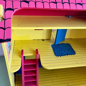Op de afbeelding: Een felgekleurd houten poppenhuis met een roze dak en gele vloeren. Het poppenhuis heeft een rode ladder, blauwe accenten en een blauw tapijt. Het dak heeft zwarte randen. Het poppenhuis is ontworpen met meerdere niveaus en kamers.