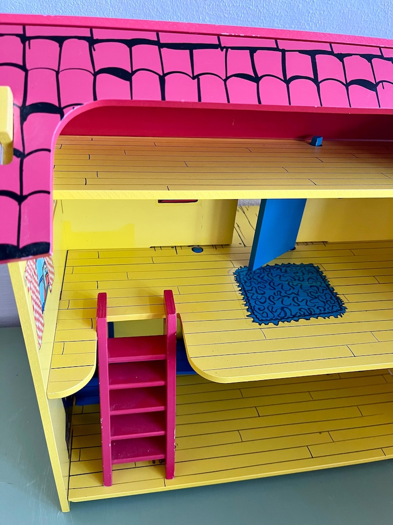 Op de afbeelding: Een felgekleurd houten poppenhuis met een roze dak en gele vloeren. Het poppenhuis heeft een rode ladder, blauwe accenten en een blauw tapijt. Het dak heeft zwarte randen. Het poppenhuis is ontworpen met meerdere niveaus en kamers.