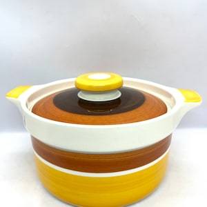 Puede incluir: Una cacerola de cerámica blanca con un diseño de rayas amarillas y marrones. La tapa tiene un pomo amarillo.