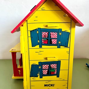 Op de afbeelding: Een geel houten speelgoedhuis met een rood dak en blauwe luiken. Het huis heeft twee ramen met rood-witte geruite gordijnen en kleine potplanten. Het woord "MICKI" staat op de voorkant.