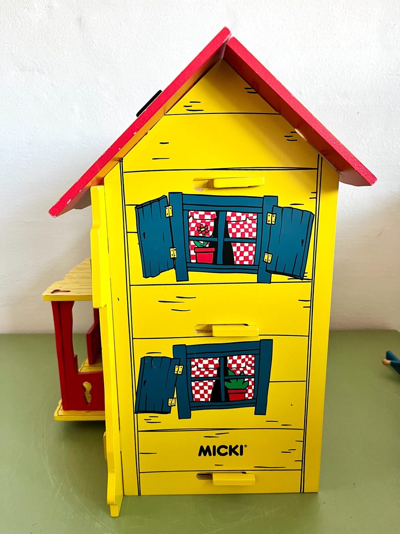 Op de afbeelding: Een geel houten speelgoedhuis met een rood dak en blauwe luiken. Het huis heeft twee ramen met rood-witte geruite gordijnen en kleine potplanten. Het woord "MICKI" staat op de voorkant.