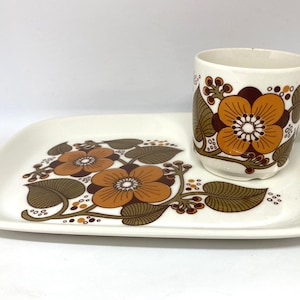 Puede incluir: Un juego de taza y bandeja rectangular de cerámica blanca. La taza y la bandeja presentan un patrón floral repetido en tonos naranja, marrón y blanco. Las flores tienen un diseño detallado con tallos y hojas marrones. Ideal para café o té.