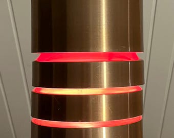 May include: A cylindrical copper-colored pendant light fixture with three illuminated red bands. The light hangs from a gray cord. The fixture has a modern design and is likely used for interior lighting.