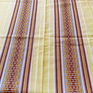 May include: A yellow tablecloth with vertical stripes in white, gold, gray, and maroon. The center of the tablecloth features a detailed geometric pattern in maroon and gold.