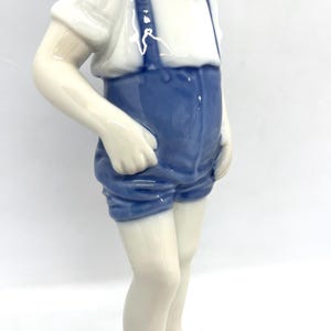 May include: A glazed porcelain figurine of a child standing upright. The figure wears a white short-sleeved shirt and blue shorts with braces. The child is depicted barefoot with hands resting near the pockets of the blue shorts.