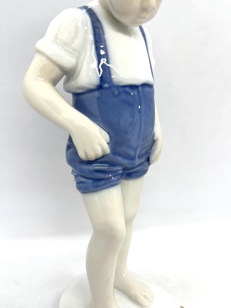 May include: A glazed porcelain figurine of a child standing upright. The figure wears a white short-sleeved shirt and blue shorts with braces. The child is depicted barefoot with hands resting near the pockets of the blue shorts.
