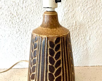 May include: A brown ceramic table lamp with a wheat stalk design. The lamp has a white socket and a black on/off switch. The lamp's base is a dark brown color with a textured surface. The lamp is plugged into an electrical outlet.