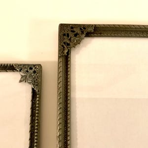May include: Two antique silver-toned picture frames with ornate corner details. The frames have a textured border and a white interior. The frames are displayed against a cream-colored background.