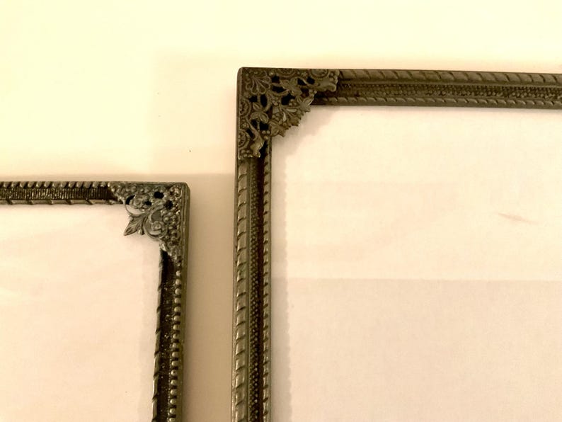 May include: Two antique silver-toned picture frames with ornate corner details. The frames have a textured border and a white interior. The frames are displayed against a cream-colored background.