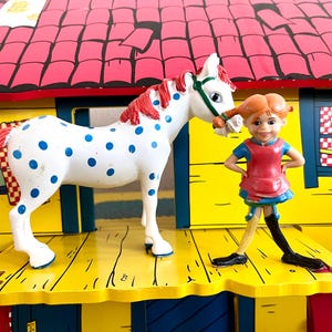 Op de afbeelding: Een wit speelgoedpaard met blauwe stippen en een rode manen en staart staat naast een beeldje van een meisje met rode vlechtjes. Het meisje draagt een rode jurk en een blauwe top. De achtergrond is een geel en rood speelhuis.
