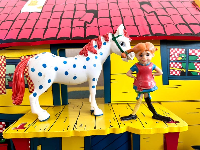 Op de afbeelding: Een wit speelgoedpaard met blauwe stippen en een rode manen en staart staat naast een beeldje van een meisje met rode vlechtjes. Het meisje draagt een rode jurk en een blauwe top. De achtergrond is een geel en rood speelhuis.