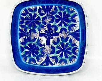 Op de afbeelding: Een vierkant keramieken bord met afgeronde hoeken voorzien van een levendig blauw en groen bloemmotief. Het ontwerp bestaat uit herhalende gestileerde blauwe bloemmotieven op een lichtgroene achtergrond, omlijst door een effen donkerblauwe rand.