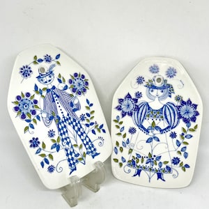 May include: Two white ceramic decorative plates with blue and green floral and figure designs. One plate features a man in a hat and striped suit, the other a woman in a dress. Both plates have a unique shape.