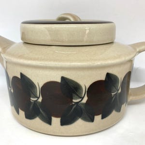 Peut inclure: Théière en céramique avec couvercle, de couleur beige avec un bord marron. La théière est décorée d'un motif répété de fruits bruns et de feuilles vertes. La poignée et le bec sont incurvés.