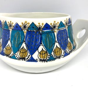 Può includere: Salsiera in ceramica bianca con un motivo di pesci blu, turchese e oro. La salsiera ha un beccuccio e un manico con un foro. Il motivo dei pesci si ripete all'esterno, creando un pezzo decorativo e funzionale.