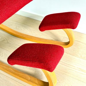 May include: A red ergonomic kneeling chair with a curved wooden frame. The chair has padded seat and knee rests, designed for a unique seating posture. The design combines wood and fabric, creating a modern aesthetic.