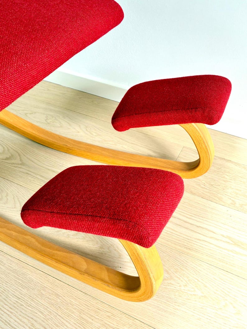 May include: A red ergonomic kneeling chair with a curved wooden frame. The chair has padded seat and knee rests, designed for a unique seating posture. The design combines wood and fabric, creating a modern aesthetic.
