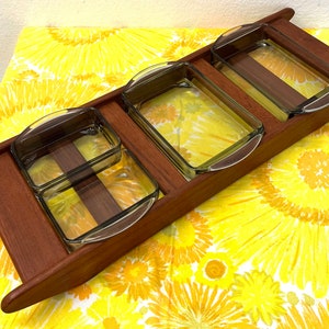 Puede incluir: Una bandeja de madera para servir con tres recipientes de vidrio transparente. La bandeja es rectangular con un acabado de madera oscura y un borde elevado. Los recipientes de vidrio son cuadrados y tienen asas curvas. La bandeja está sobre una superficie amarilla con estampado floral.