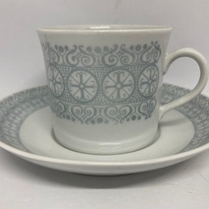 Może przedstawiać: Biały ceramiczny kubek do kawy i spodek z geometrycznym wzorem w kolorze szarym i białym. Kubek ma uchwyt, a spodek ma falisty brzeg.