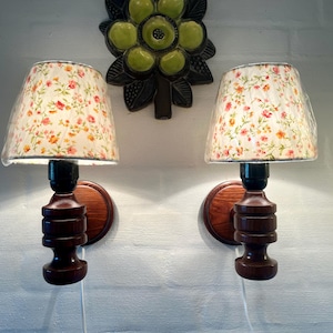 Może przedstawiać: Para w stylu vintage kinkietów z kwiatowymi abażurami. Lampy mają ciemnobrązowe drewniane podstawy i okrągłe mocowania ścienne. Abażury mają białe tło z czerwonymi, pomarańczowymi i żółtymi kwiatami. Dekoracja ścienna znajduje się nad lampami.